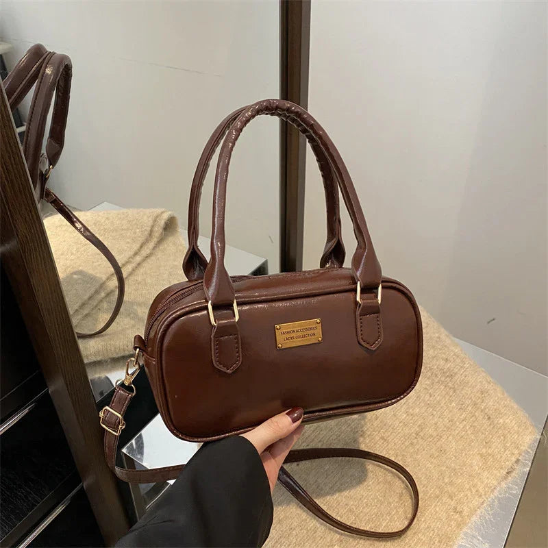 Brown leather handbag with dual handles and gold plaque, held over beige surface, near mirror