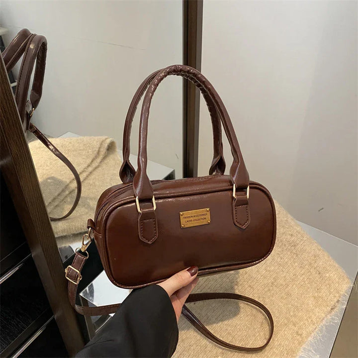 Brown leather handbag with dual handles and gold plaque, held over beige surface, near mirror