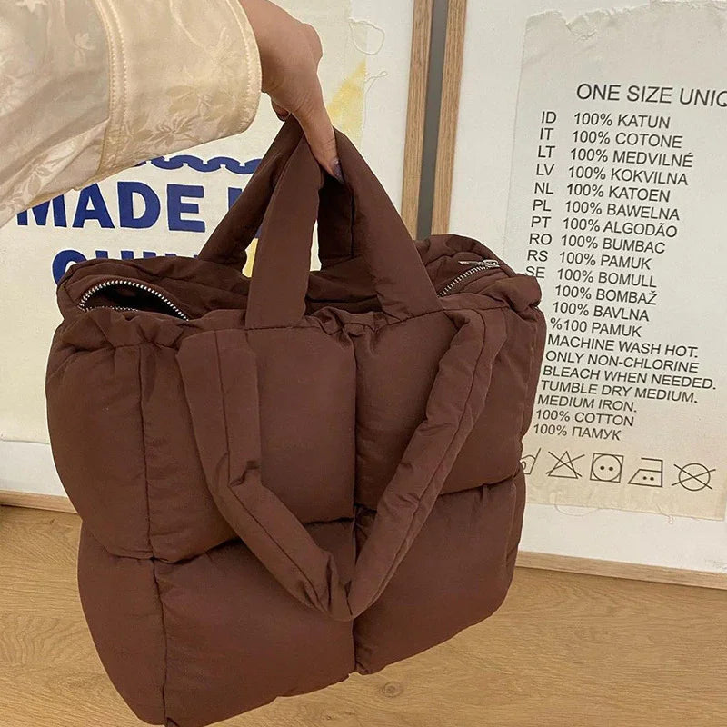 Hand holding a brown padded tote bag with zippered pockets on wooden floor backdrop