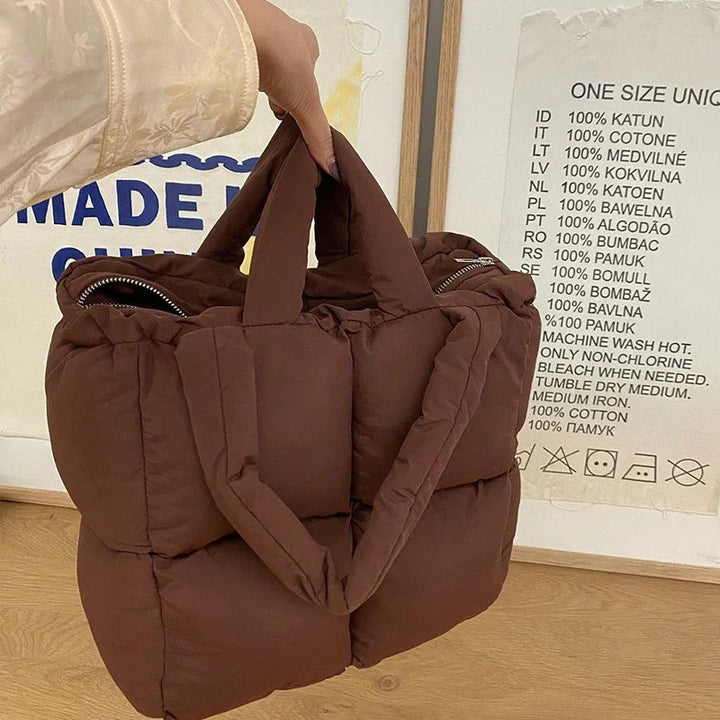 Hand holding a brown padded tote bag with zippered pockets on wooden floor backdrop
