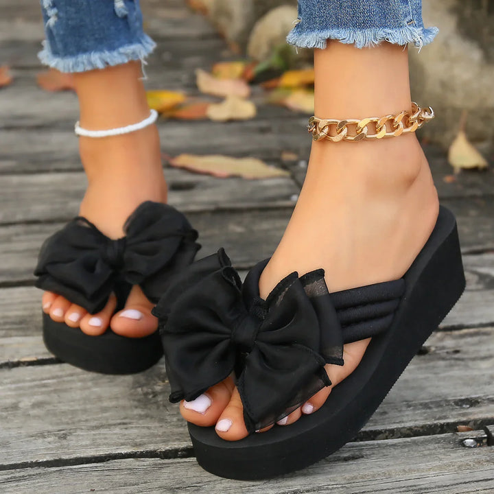 Woman wearing black platform sandals with large bows and gold and white ankle bracelets on wooden floor