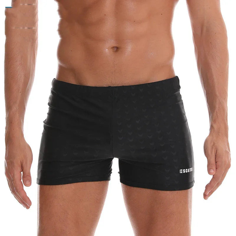 Close-up of a muscular man wearing black square-cut swim trunks with subtle heart patterns and ESCAPE logo