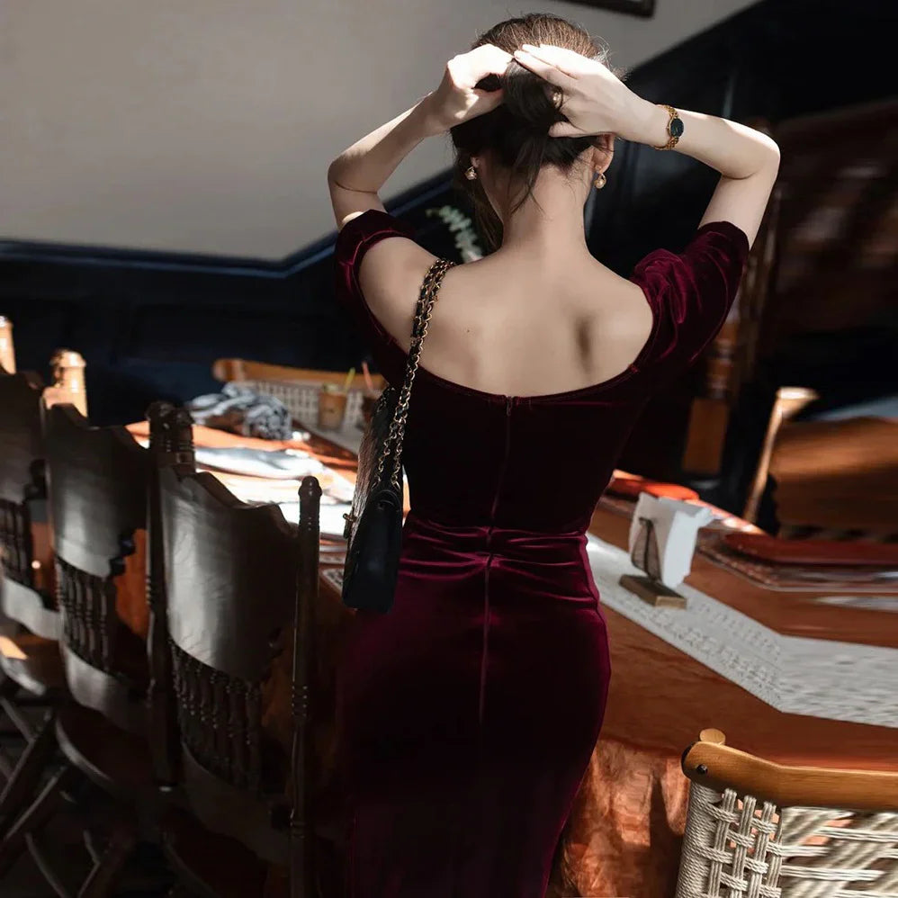 Woman in a dark red velvet dress adjusting hair, standing by a wooden dining table with chairs
