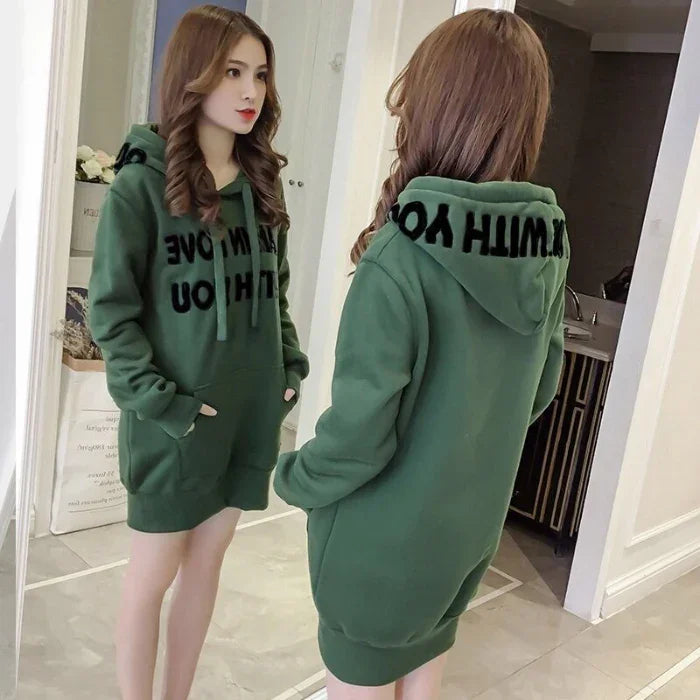Young woman wearing a green oversized hoodie dress with black text, standing in front of a mirror indoors