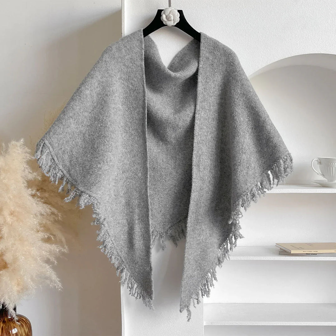 Soft gray fringed wool poncho on black hanger against white wall with bookshelf and pampas grass