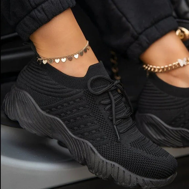 Close-up of black knitted sneakers with thick soles worn with black joggers and gold heart charm anklet