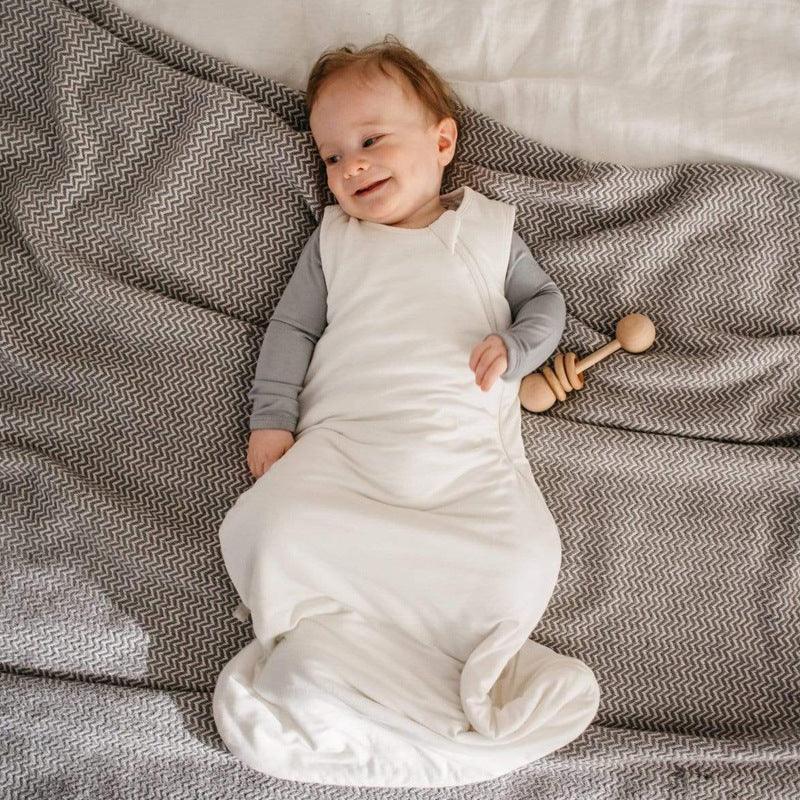 Smiling baby lying on a grey zigzag blanket, wearing a white sleep sack over grey clothing, holding a wooden rattle