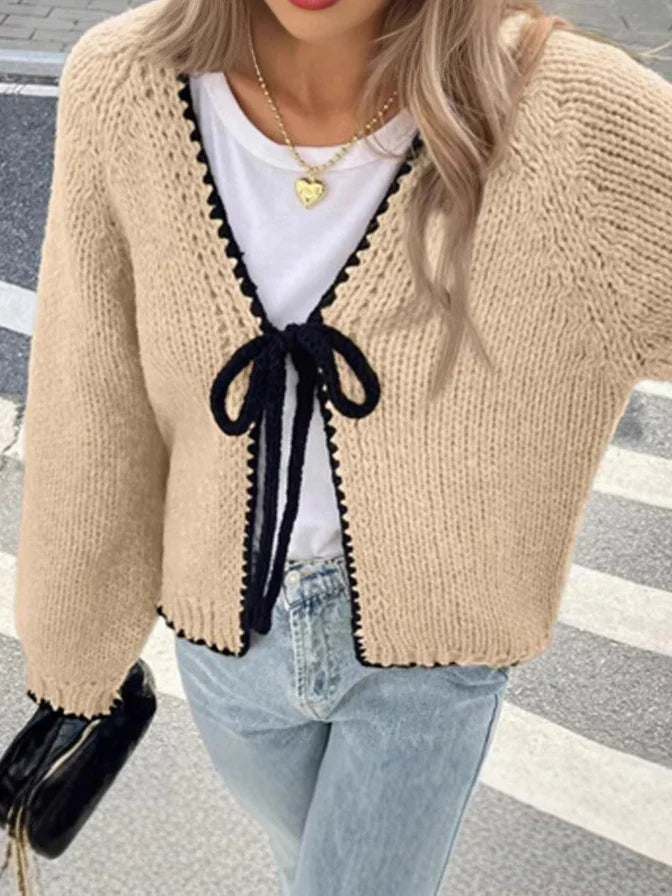 Woman wearing beige knit cardigan with black trim and bow over white top and light blue jeans on street