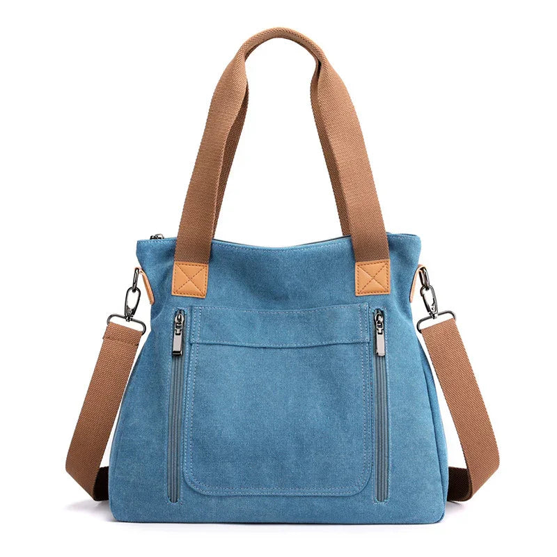 Blue canvas tote bag with brown handles and adjustable shoulder strap, front zip pockets