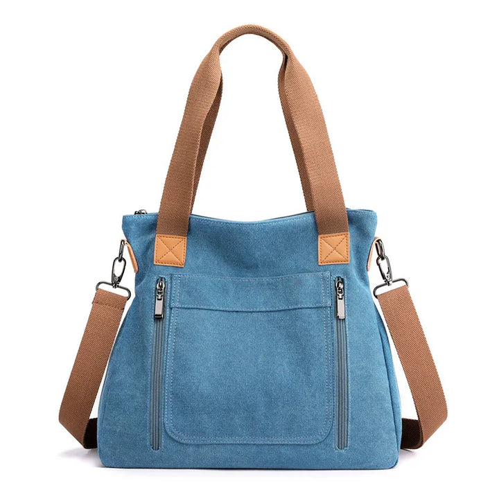 Blue canvas tote bag with brown handles and adjustable shoulder strap, front zip pockets