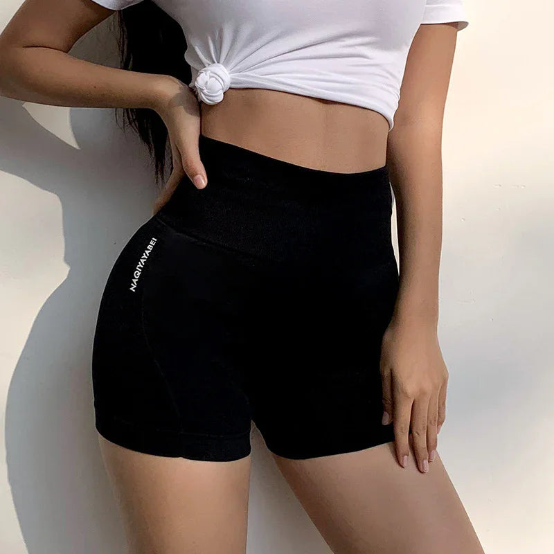 Woman wearing high-waisted black workout shorts and a knotted white crop top against plain wall