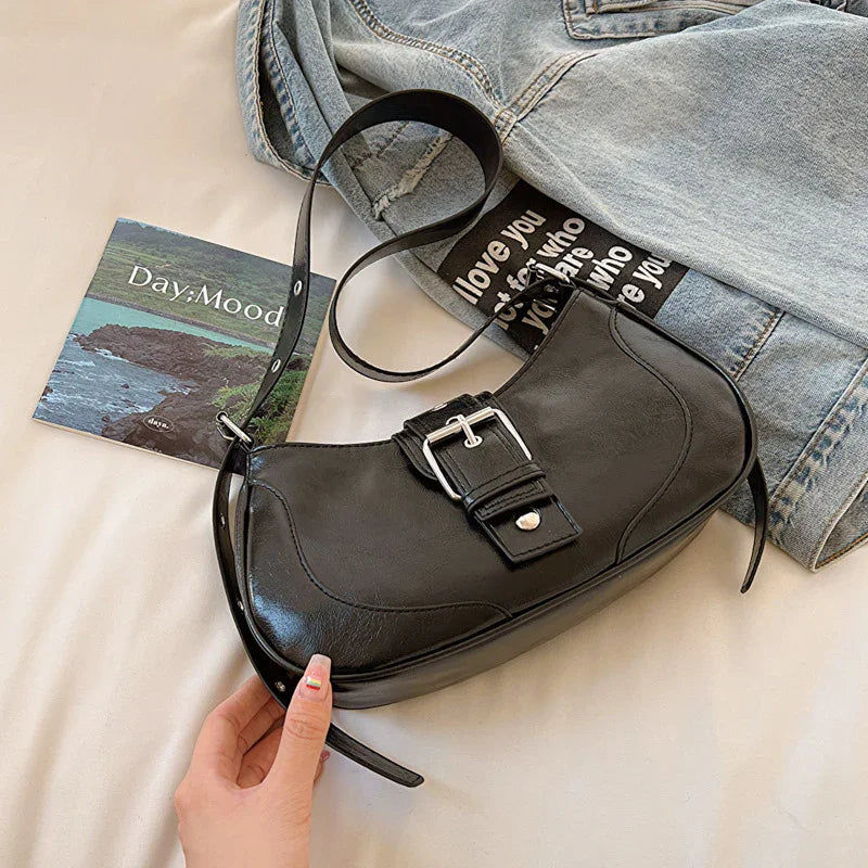 Black leather shoulder bag with silver buckle on bed with denim jacket and scenic postcard