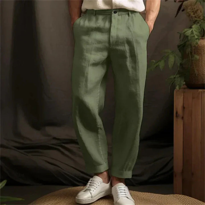 Man wearing olive green casual pants and white sneakers against dark backdrop with plant decor