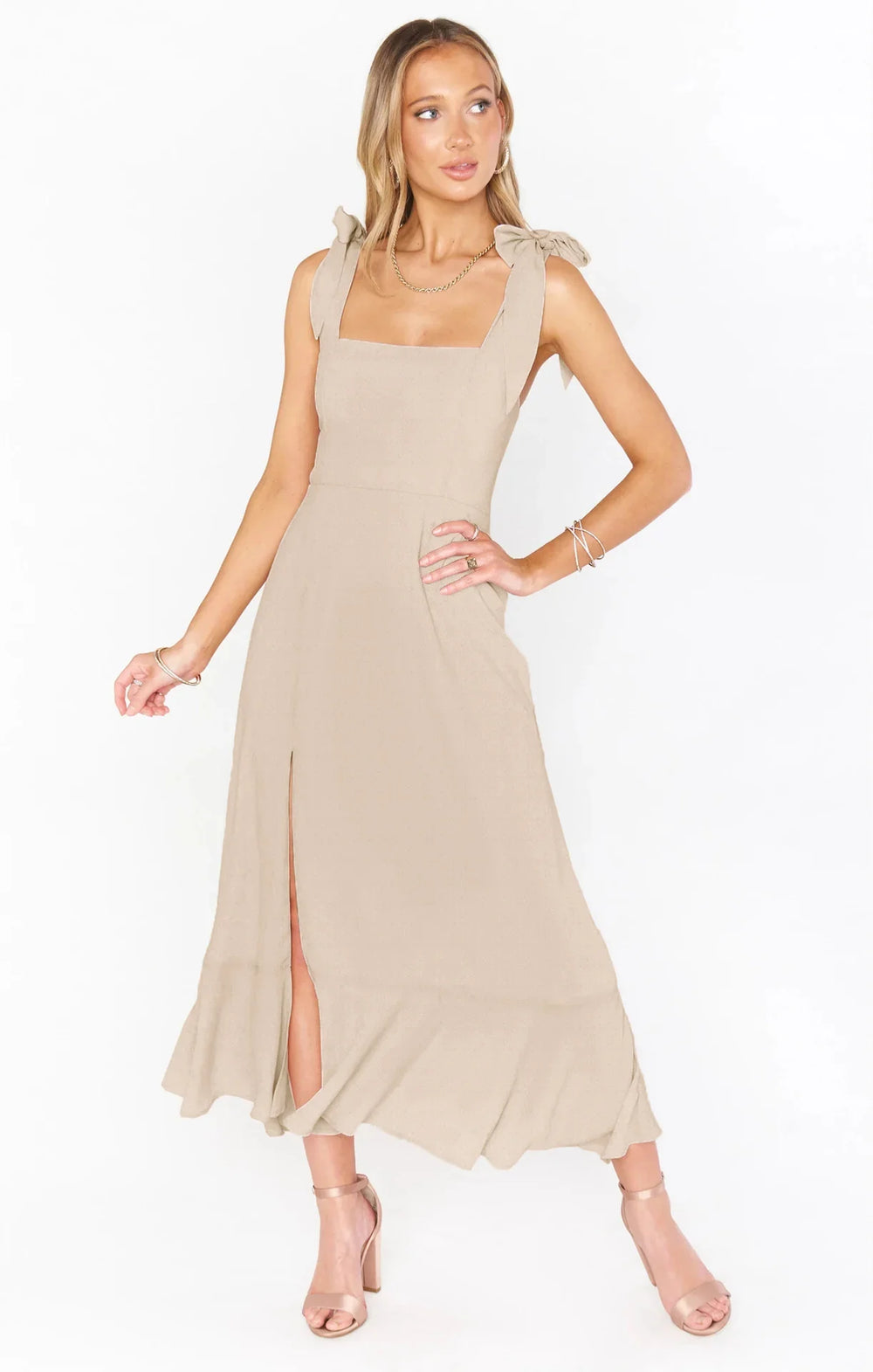 Woman wearing beige midi dress with bow shoulder straps and front slit, paired with nude heels