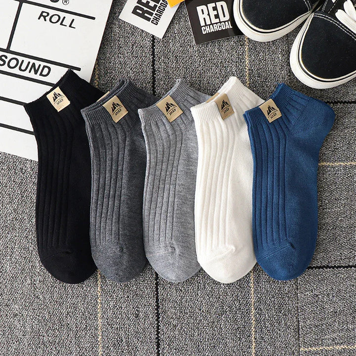 Set of five ribbed ankle socks in black, dark gray, light gray, white, and blue on gray carpet with sneakers and paper tags