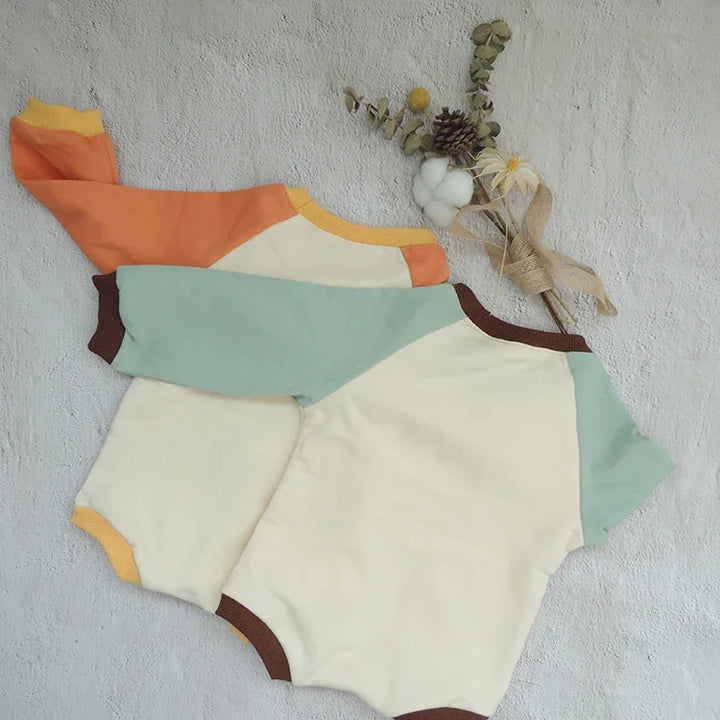 Two baby rompers with raglan sleeves in cream with orange and mint, styled with dried flowers on grey fabric