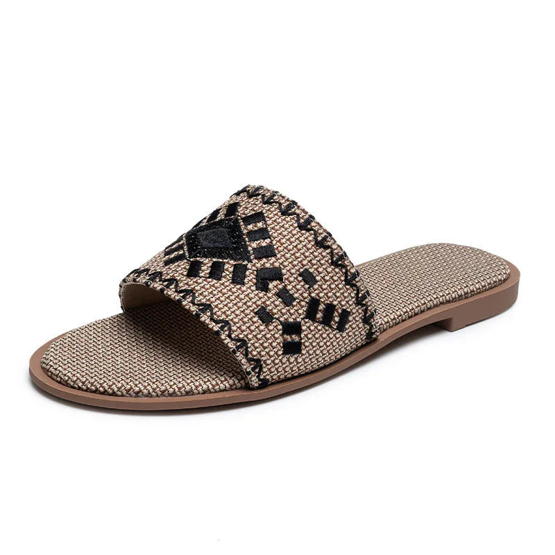 Beige woven slide sandal with black geometric embroidery and flat sole on white background