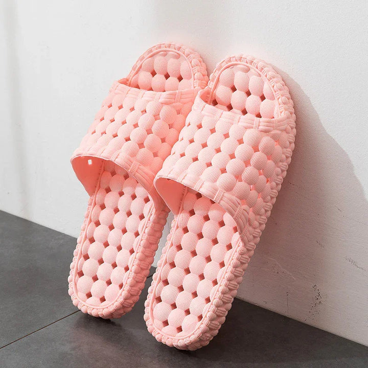 Pair of pink textured open-toe slippers with raised circular pattern on gray floor against white wall