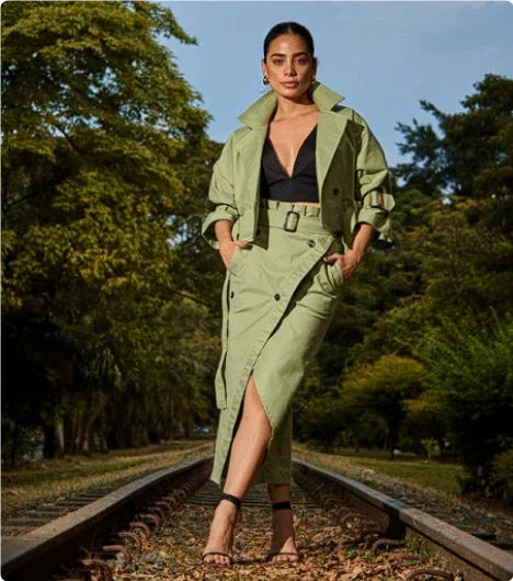 Woman in stylish sage green trench coat and skirt walking confidently on railway tracks surrounded by trees