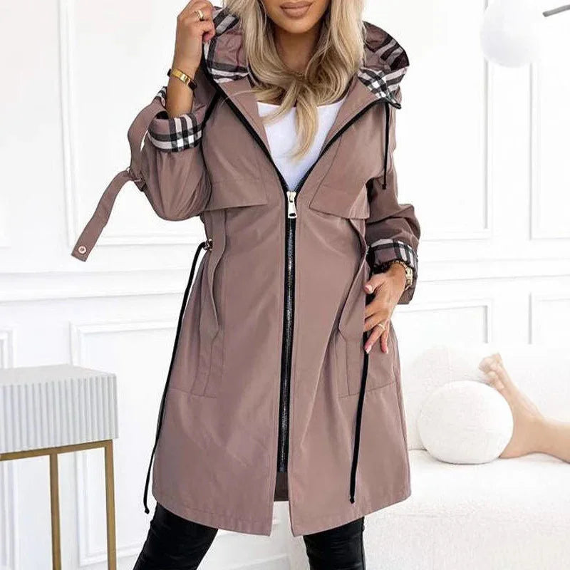 Woman wearing a light brown hooded trench coat with plaid cuffs and zipper in a white room