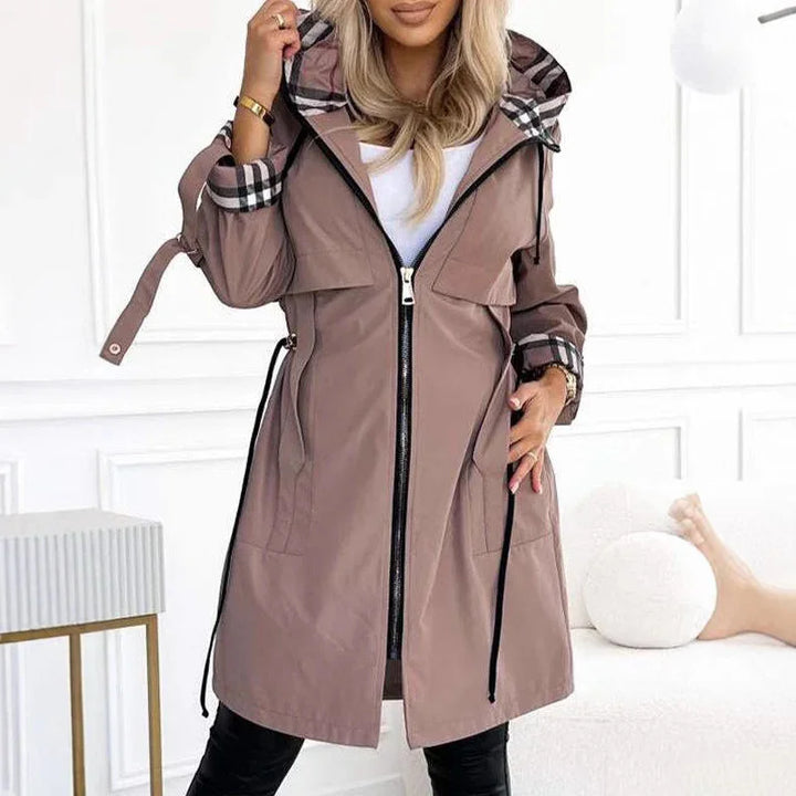 Woman wearing a light brown hooded trench coat with plaid cuffs and zipper in a white room