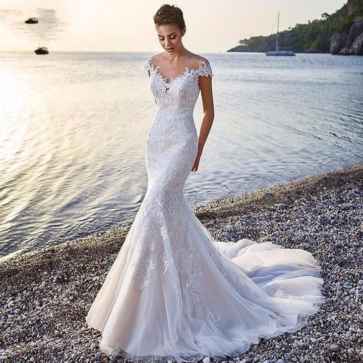 Bride in elegant lace mermaid wedding dress standing on pebble beach at sunset with sailboats on sea