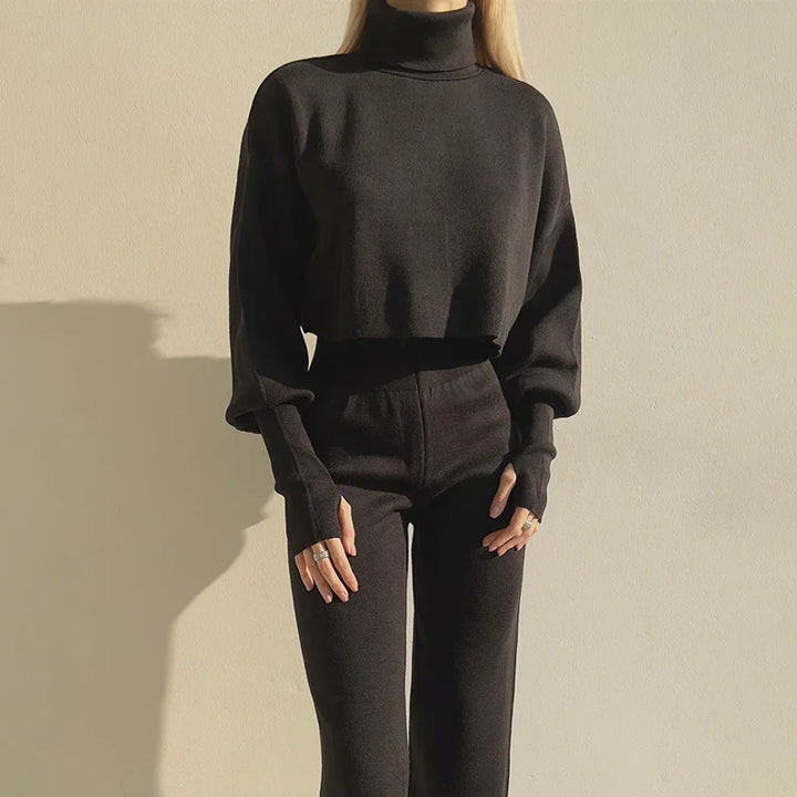 Woman wearing a black turtleneck cropped sweater and high-waisted black pants against a beige wall