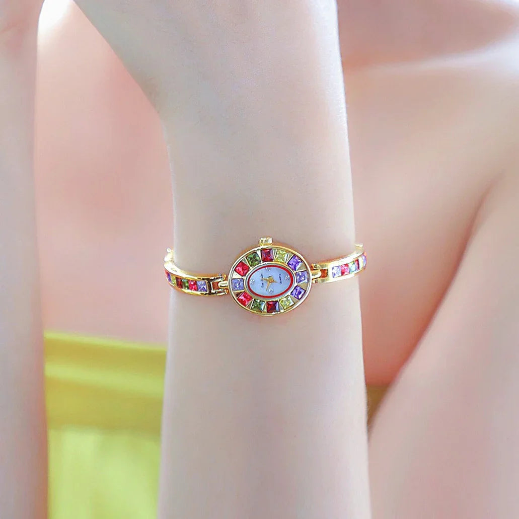 Gold bracelet watch with colorful gemstones on wrist against soft pink and yellow background