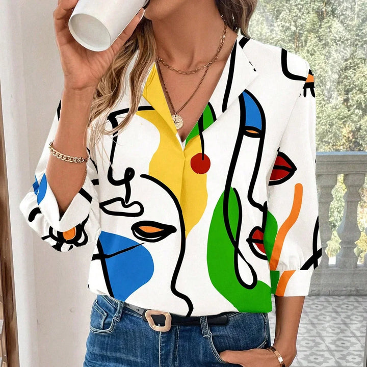 Woman wearing colorful abstract face print blouse with gold necklace and jeans, drinking from white cup