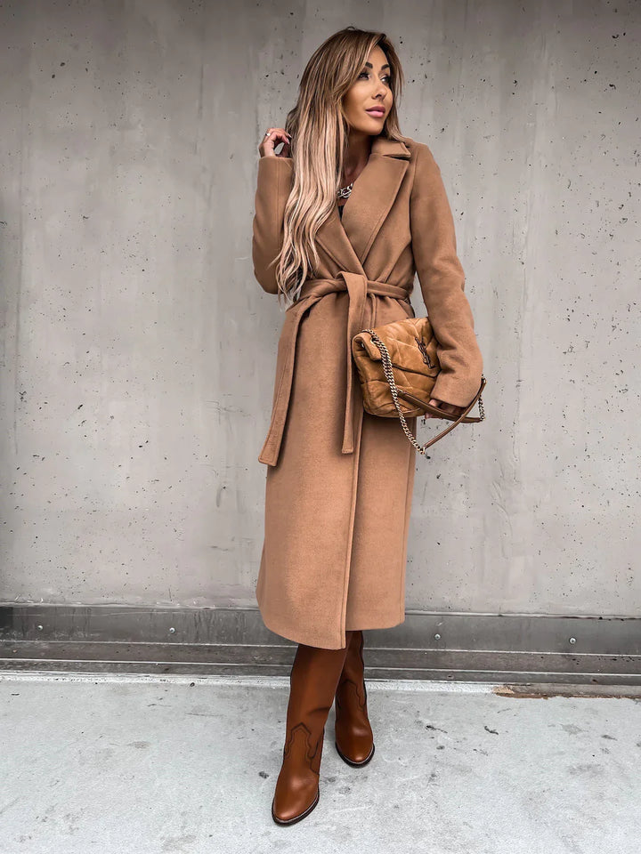 woman wearing long camel wrap coat with belt, brown leather boots, and tan quilted chain bag against grey wall