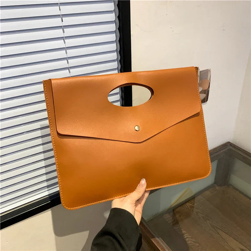 Hand holding a sleek brown leather envelope clutch bag with a single snap button closure near window blinds
