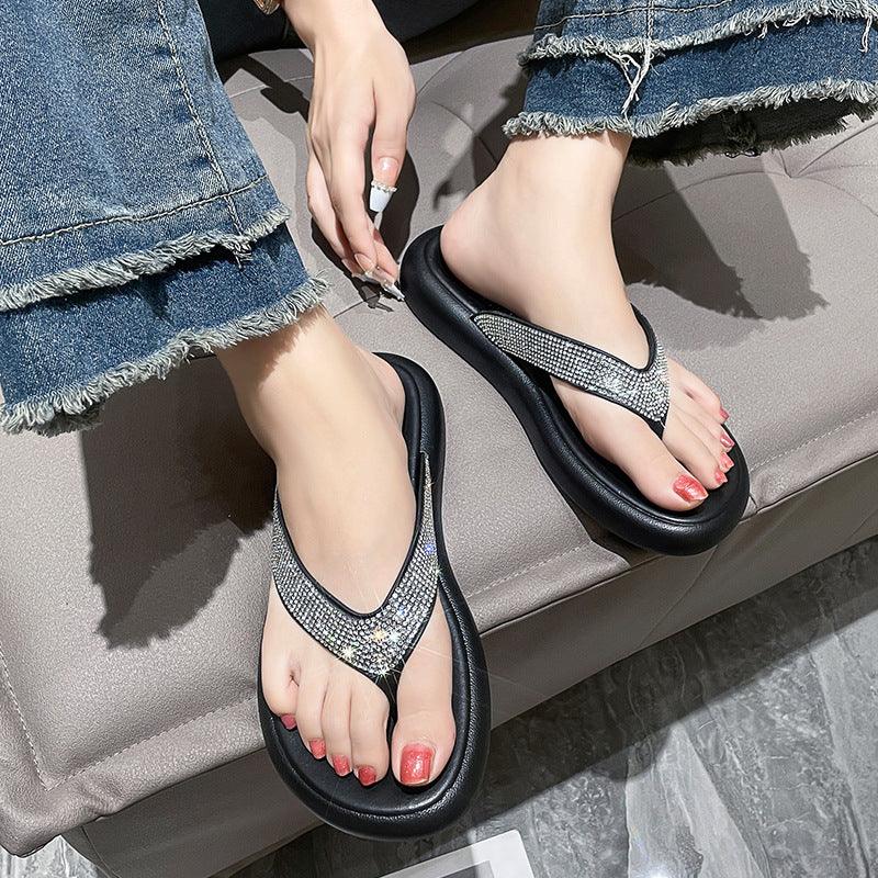 Woman wearing black cushioned flip-flops with rhinestone straps and red painted toenails, seated on gray sofa