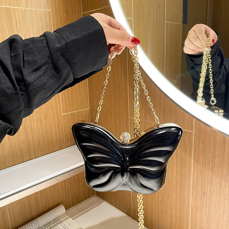 Hand holding black butterfly-shaped handbag with gold chain strap near mirror in wooden interior