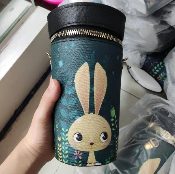 Hand holding a cylindrical teal handbag with a cartoon bunny and floral design, gold zipper