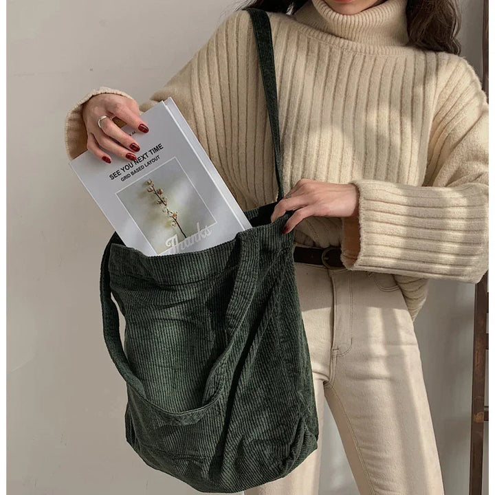 Woman in beige ribbed turtleneck sweater and cream pants putting magazine into dark green corduroy tote bag
