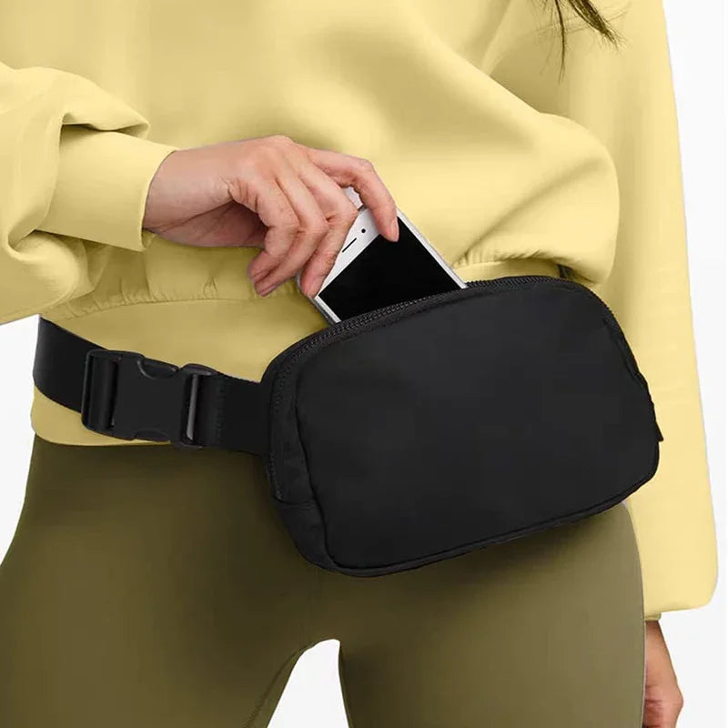 Person wearing yellow sweatshirt and olive pants placing smartphone into black waist bag fanny pack