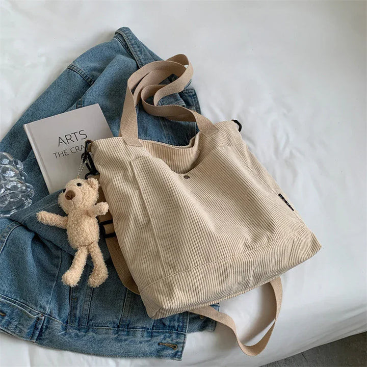 Beige corduroy tote bag with teddy bear keychain on blue denim jacket beside art book on white surface