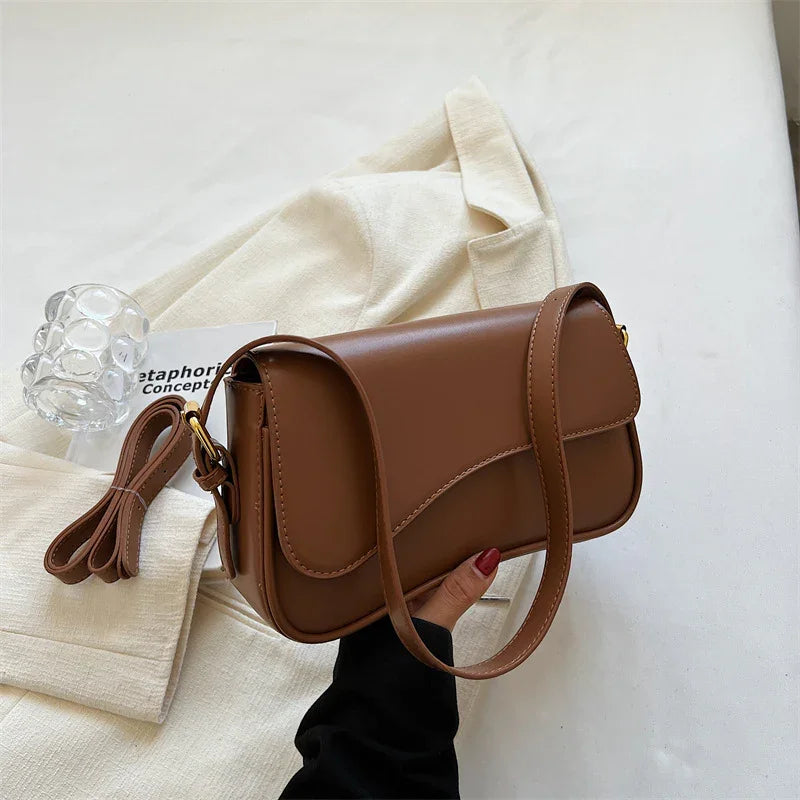 Hand holding a brown leather crossbody bag on a cream fabric background with an extra strap and clear glass decor