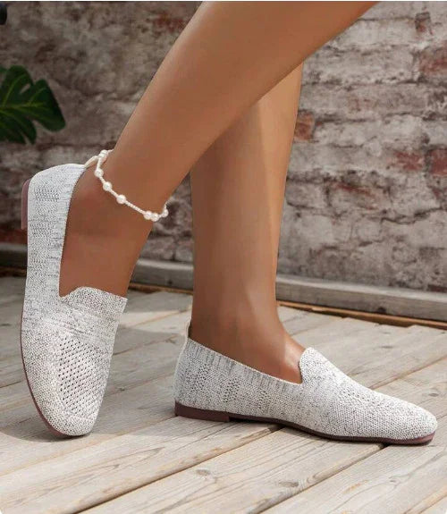 Close-up of feet wearing light gray knitted slip-on shoes and a pearl anklet on wooden floor