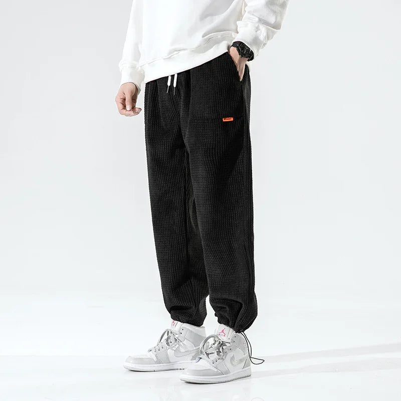Person wearing black corduroy jogger pants with drawstring ankles and white sneakers on a white background
