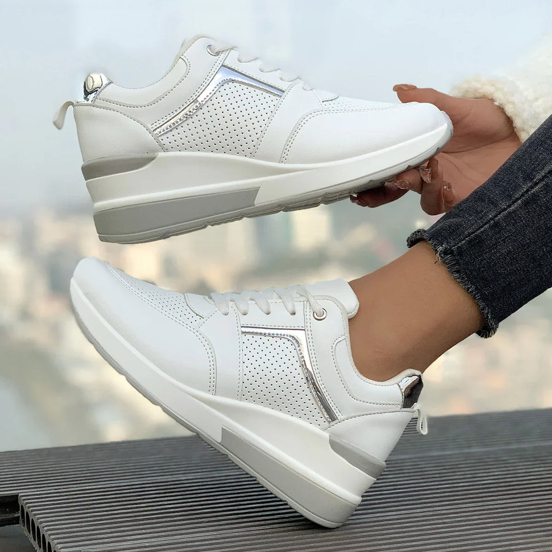 Pair of white perforated leather sneakers with silver accents worn on feet against blurred cityscape background