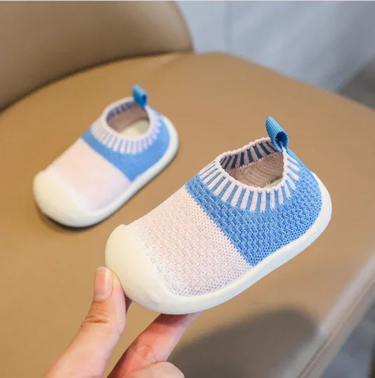 Hand holding soft knit baby shoes in pastel pink and blue with white soles on beige chair