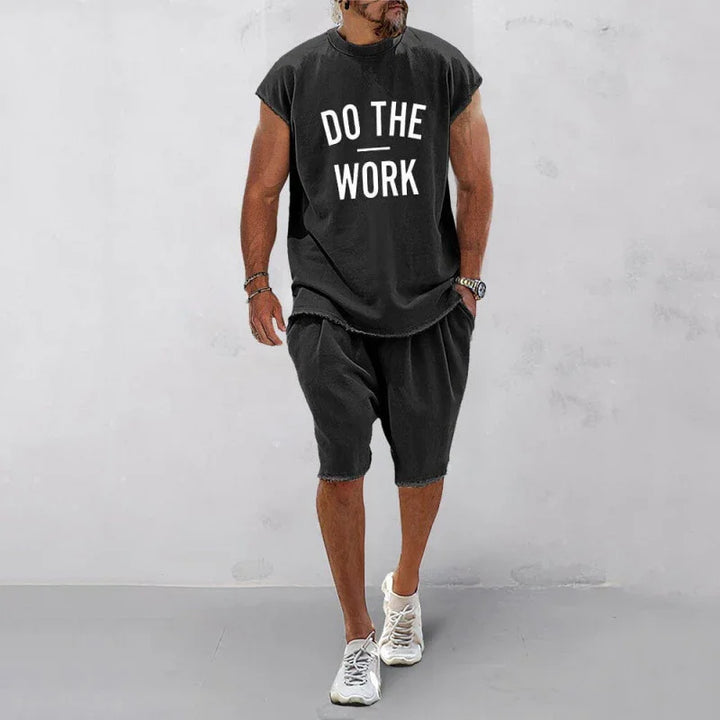 man wearing black sleeveless shirt and shorts with Do The Work text, white sneakers against gray wall