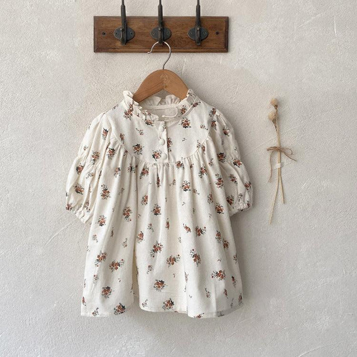 White baby dress with small floral print hanging on wooden hook rack against textured wall