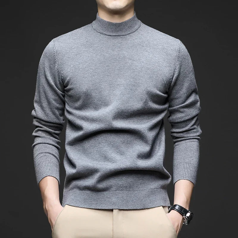 Man wearing a fitted gray turtleneck sweater with beige pants and a wristwatch on black background