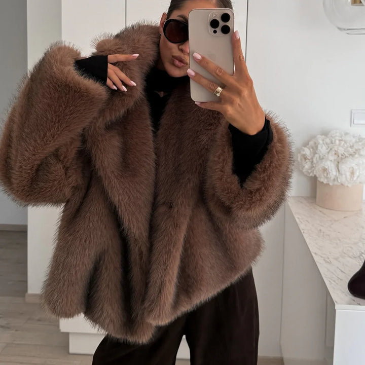 Woman wearing oversized brown faux fur coat and black sunglasses taking mirror selfie indoors