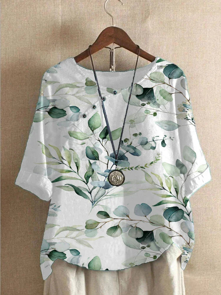 White blouse with green watercolor leaf print and round neckline displayed on wooden hanger with pendant necklace