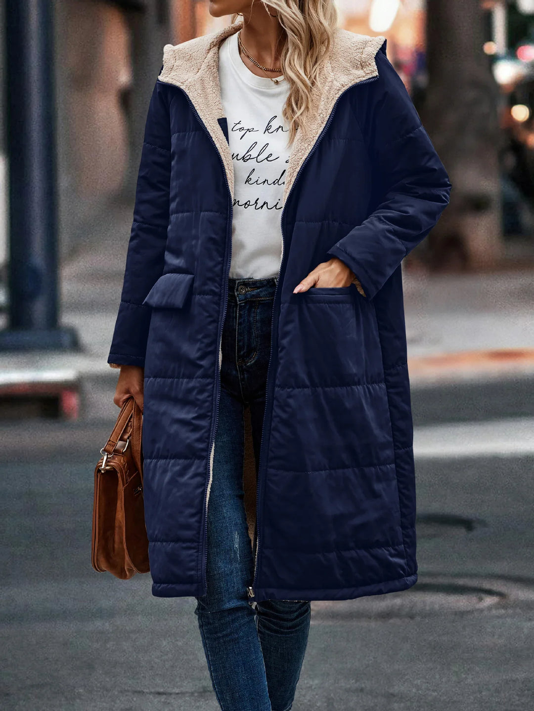 Woman wearing navy blue quilted long coat with beige sherpa lining, paired with jeans and carrying brown bag