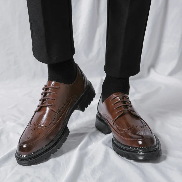 Brown leather brogue dress shoes with black soles worn with black pants and socks on white background