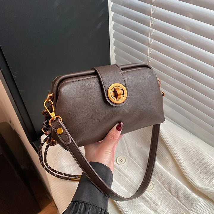 Hand holding a dark brown leather crossbody bag with gold turn-lock clasp near window blinds